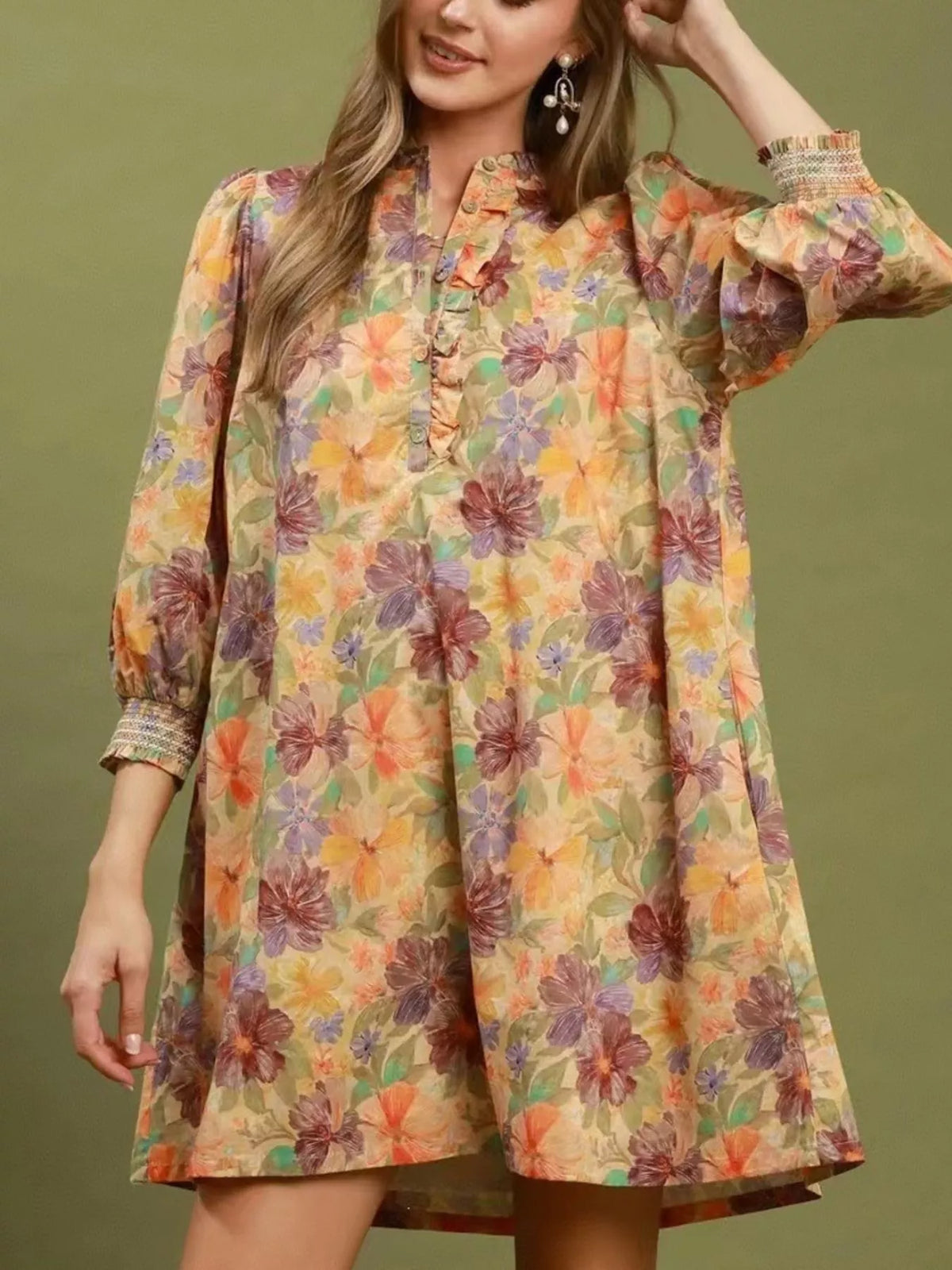 A woman wearing a long-sleeve floral tunic dress with a high ruffled collar and puff sleeves, set against a solid olive green background.
