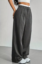 A person wearing loose, wide-leg gray pinstriped pants with a white waistband, paired with a white crop top and white sneakers, standing against a light gray wall.