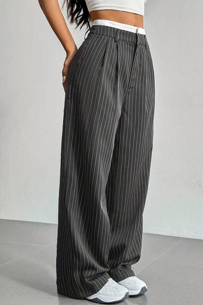 A person wearing loose, wide-leg gray pinstriped pants with a white waistband, paired with a white crop top and white sneakers, standing against a light gray wall.