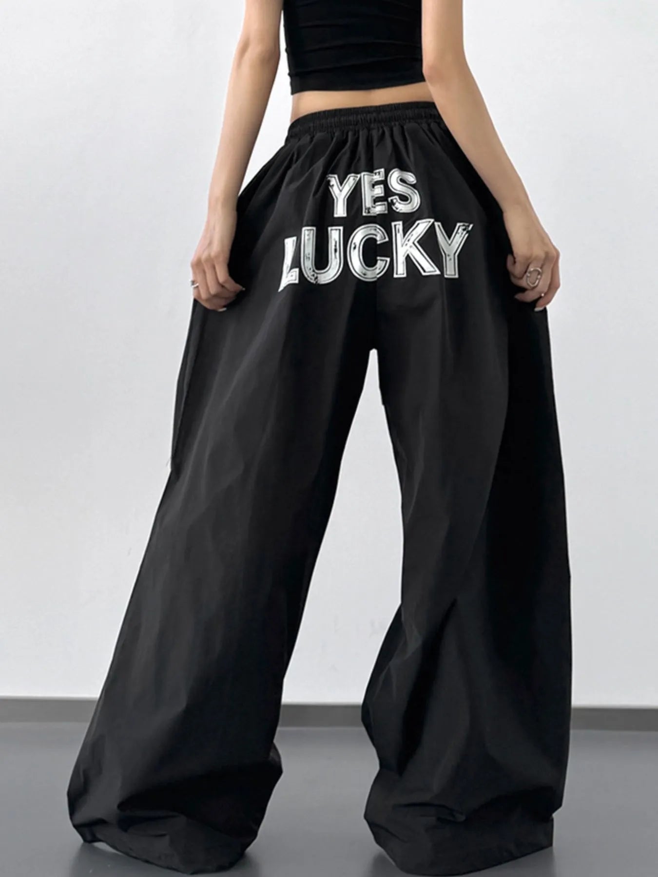 Black wide-leg pants with 'YES LUCKY' text worn by a person on a plain background