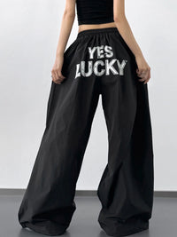 Black wide-leg pants with 'YES LUCKY' text worn by a person on a plain background