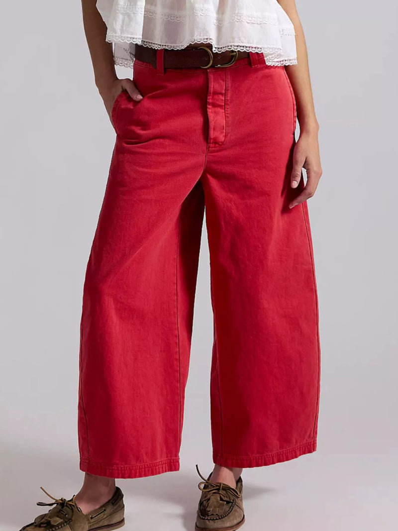 A person wearing bright red wide-leg cropped pants with a brown belt, a white ruffled top, and brown boat shoes, photographed against a light gray studio background.