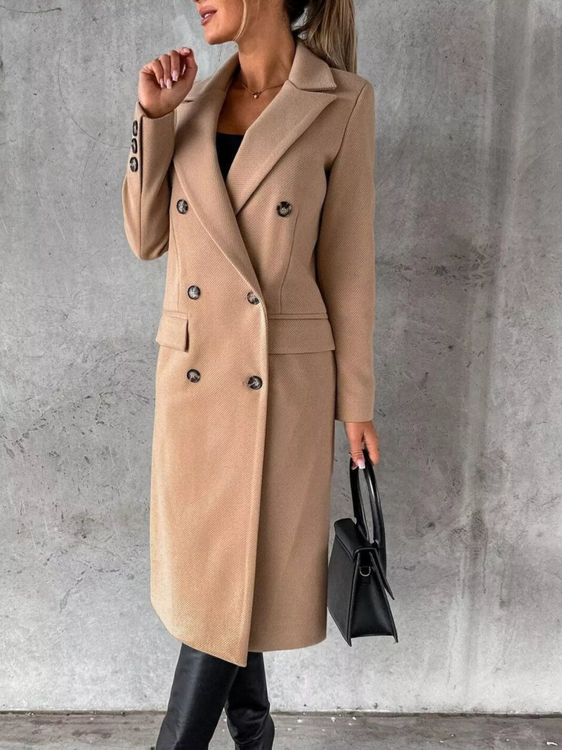 Person wearing a beige coat holding a black handbag against a gray concrete wall.