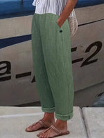 A person wearing loose sage green textured cropped pants with rolled cuffs, paired with a white striped top and brown sandals, standing in front of a boat.