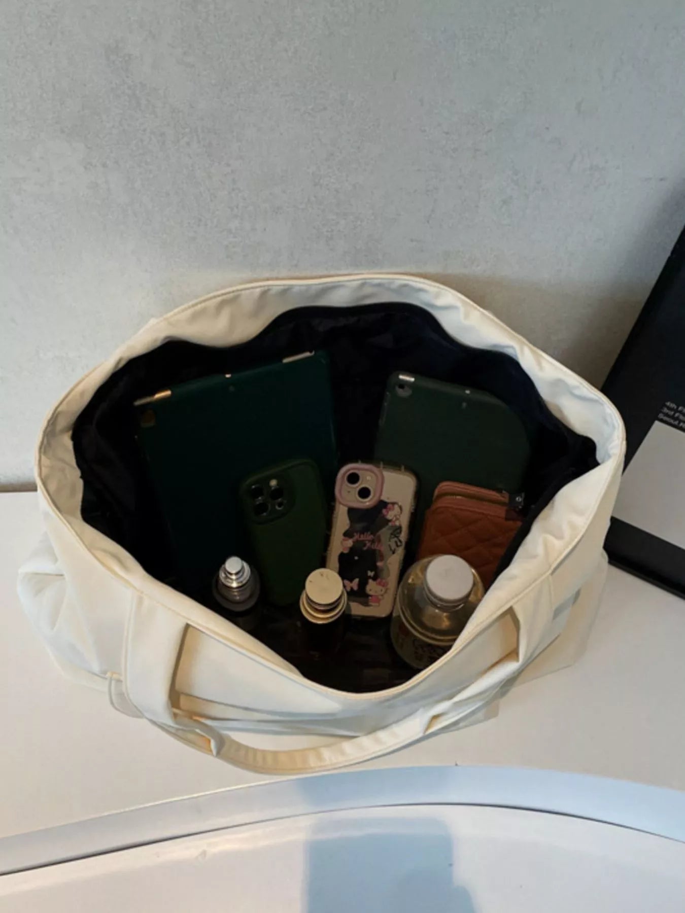 Open beige bag with electronic devices and small items on a light gray surface.