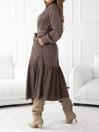 Woman wearing a long brown dress with knee-high boots in a minimalistic room.