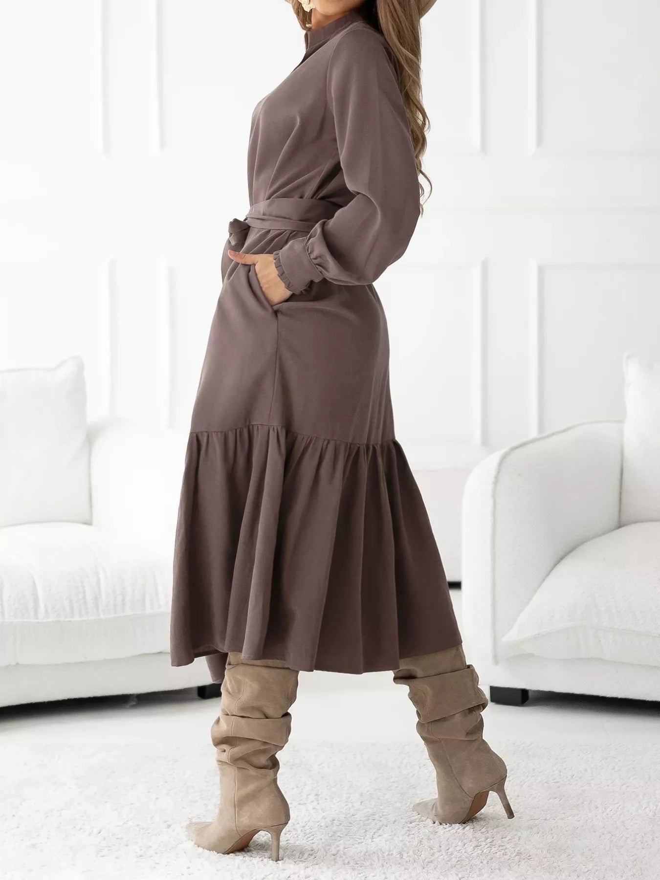 Woman wearing a long brown dress with knee-high boots in a minimalistic room.