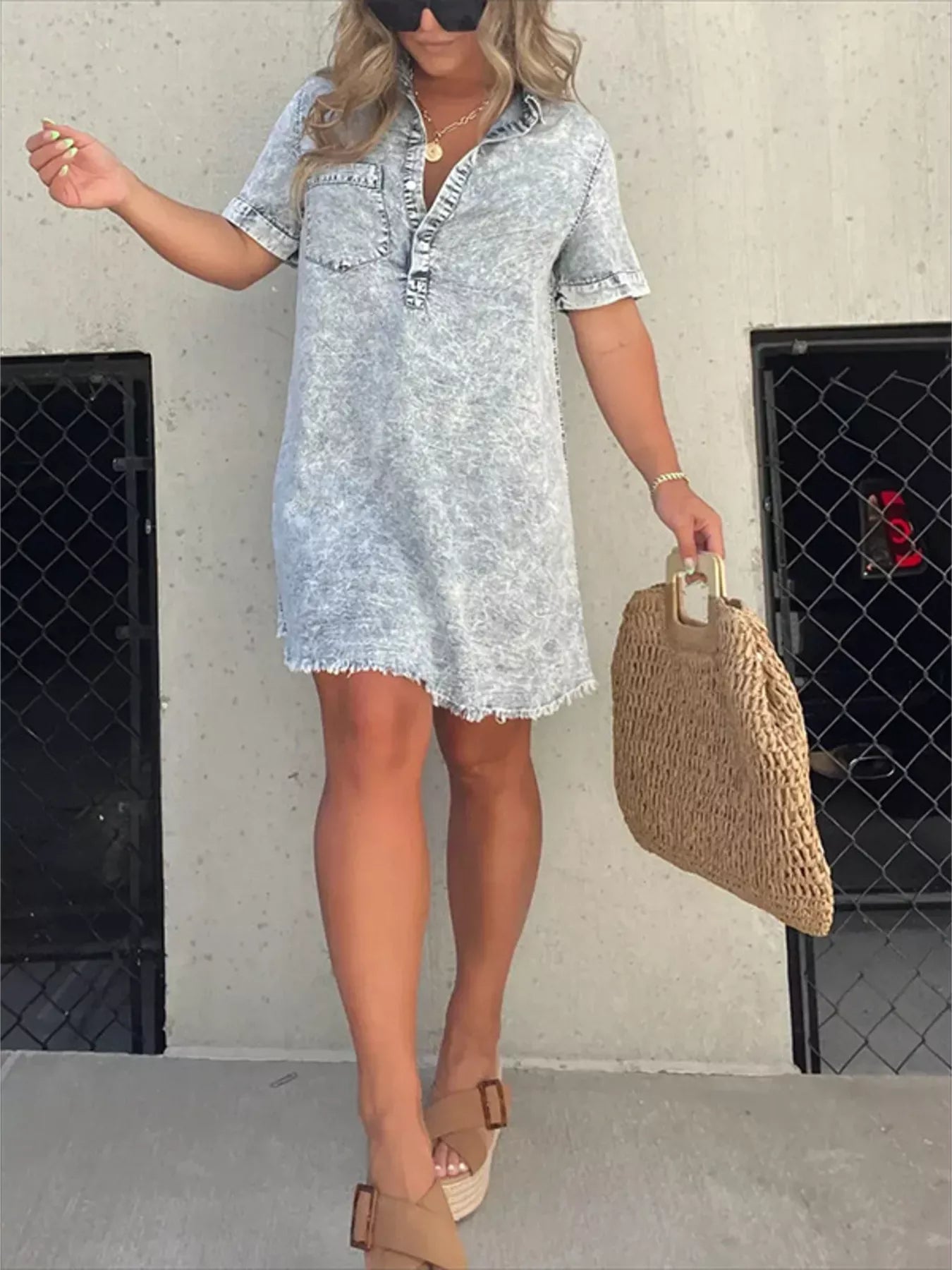 Woman wearing a light blue denim dress holding a woven bag against a plain background