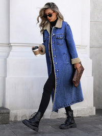 Woman wearing a long denim coat with fur trim, holding a coffee cup and clutch, standing against a white wall.