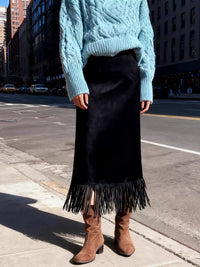 Person wearing a blue cable knit sweater, black fringed skirt, and brown boots on a city street.