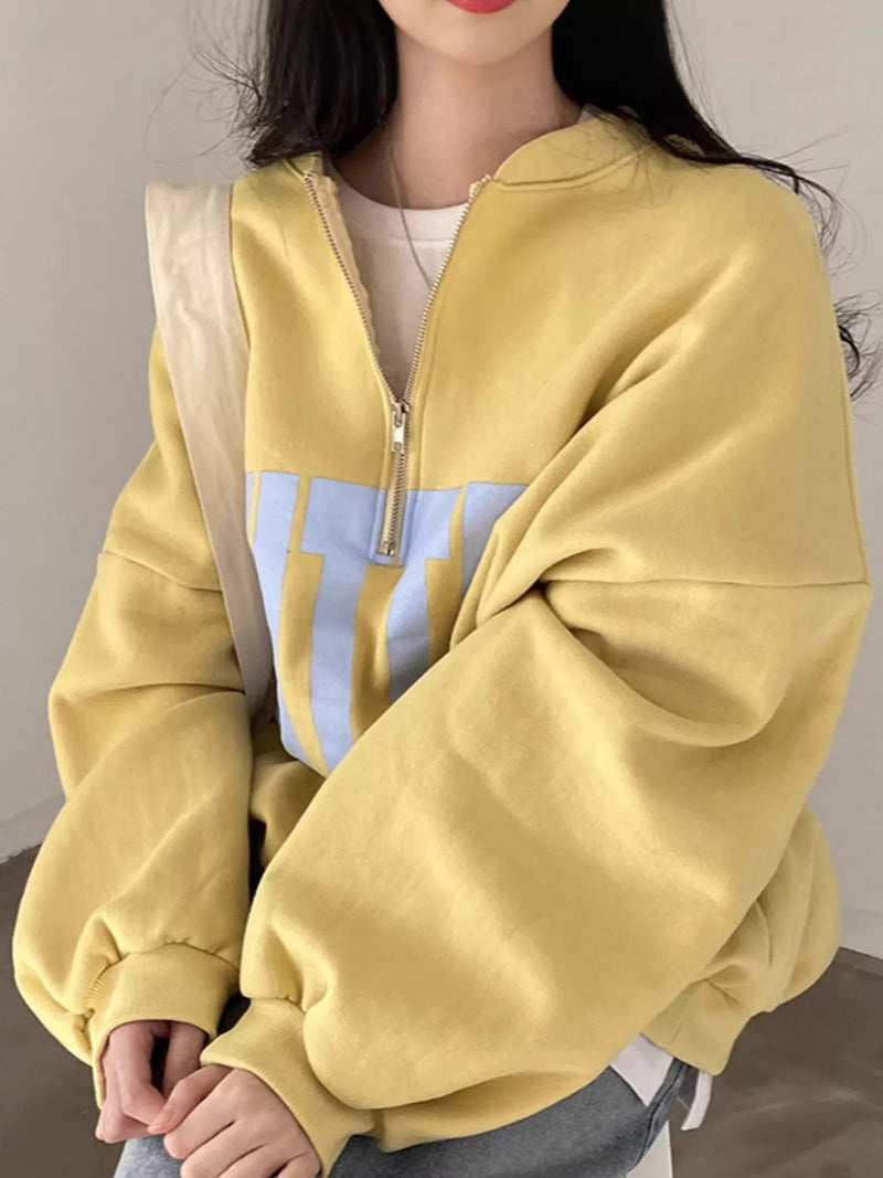 A person wearing a yellow quarter-zip sweatshirt with a light blue graphic panel on the front, layered over a white top and paired with gray pants.