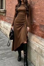 A woman wearing a brown long-sleeve midi dress with ruching and an asymmetrical hem, paired with black knee-high boots and holding a black handbag, standing against a red brick wall.