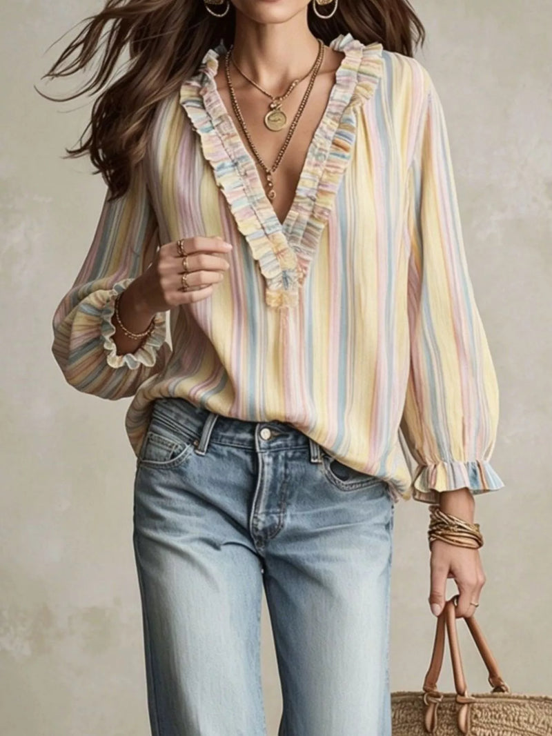 A woman wearing a pastel striped long-sleeve blouse with ruffled trim and light-wash jeans, standing against a textured beige wall and holding a woven handbag.