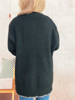 Person wearing a dark green cardigan in a room with a chair and decorative items.