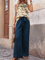 Woman wearing a floral top and blue wide-leg pants standing against a stone wall.