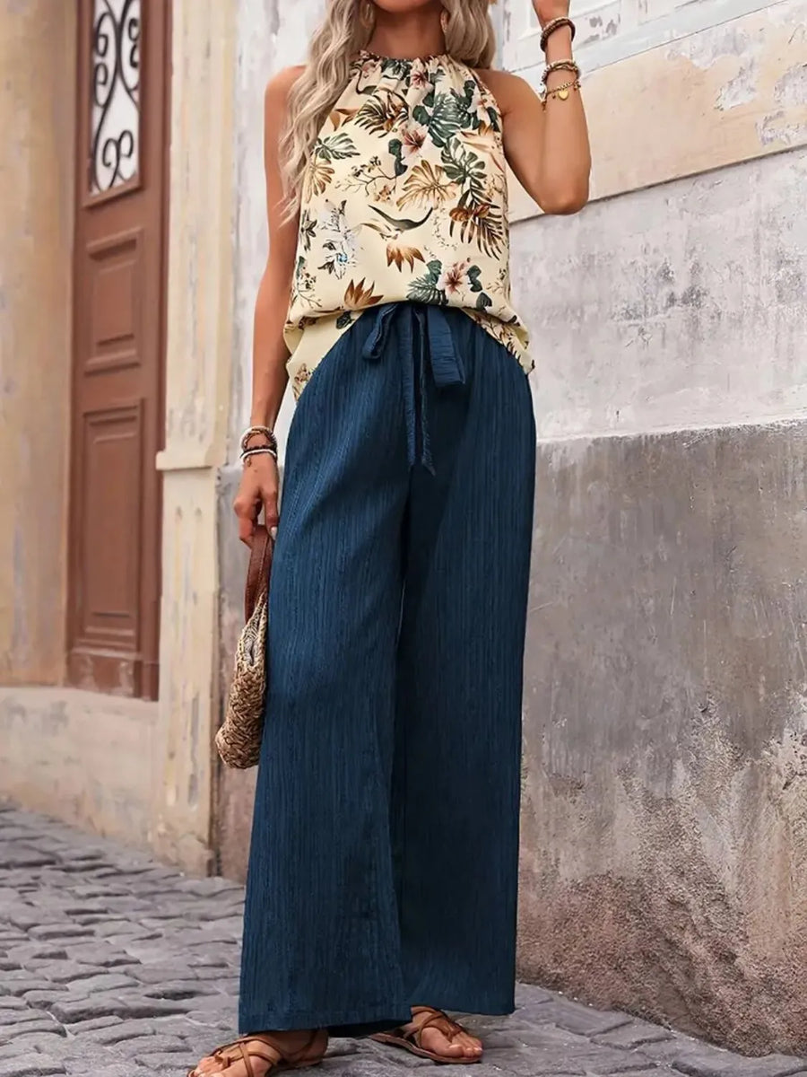 Woman wearing a floral top and blue wide-leg pants standing against a stone wall.