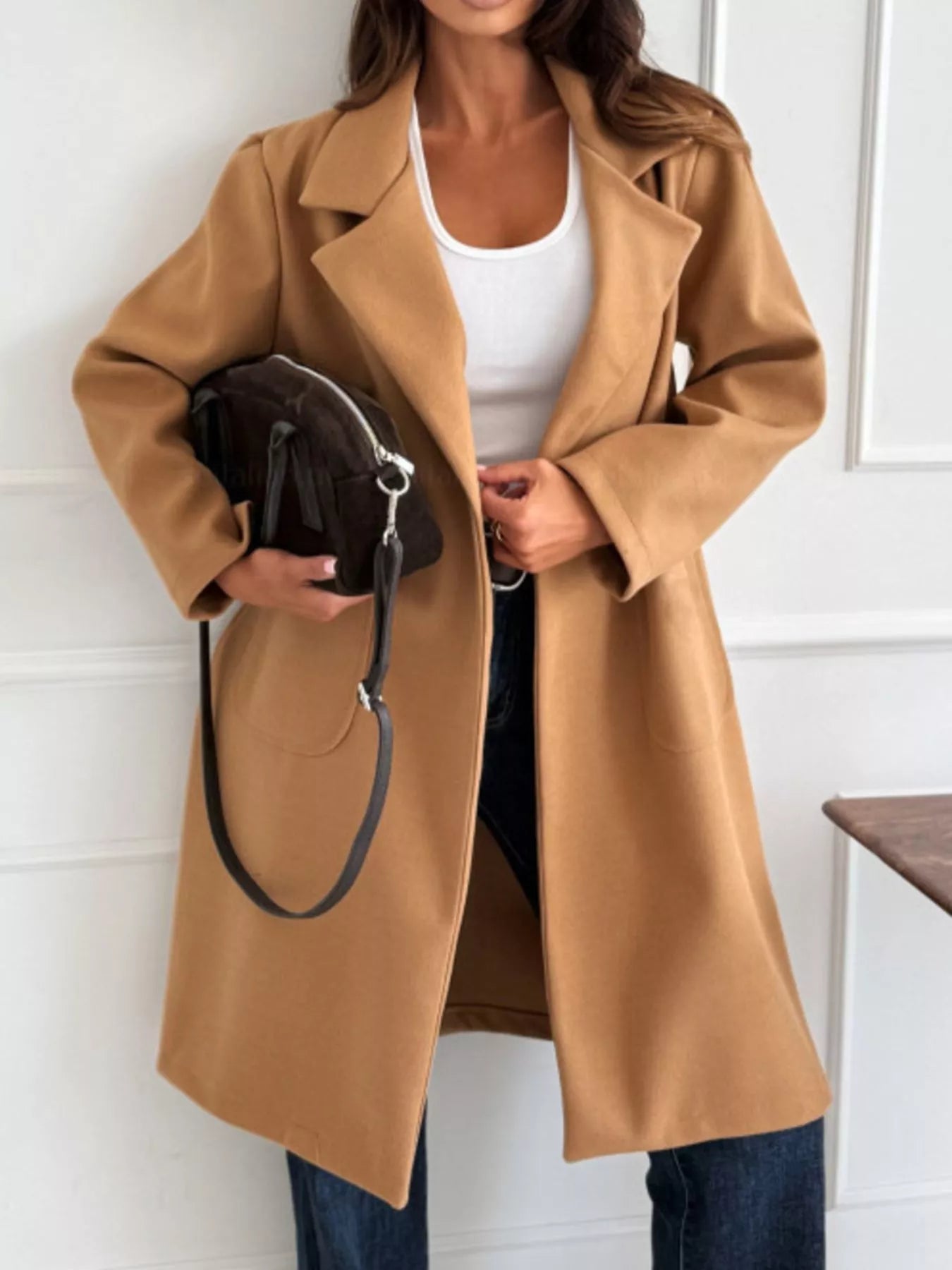 Person wearing a tan coat holding a black handbag indoors.