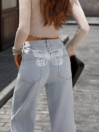 Person wearing light blue jeans with a butterfly design, standing on a street.
