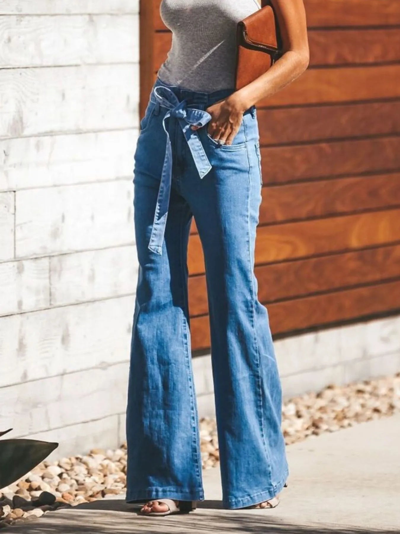 Person wearing blue flared jeans with a belt, standing against a wooden and stone wall.