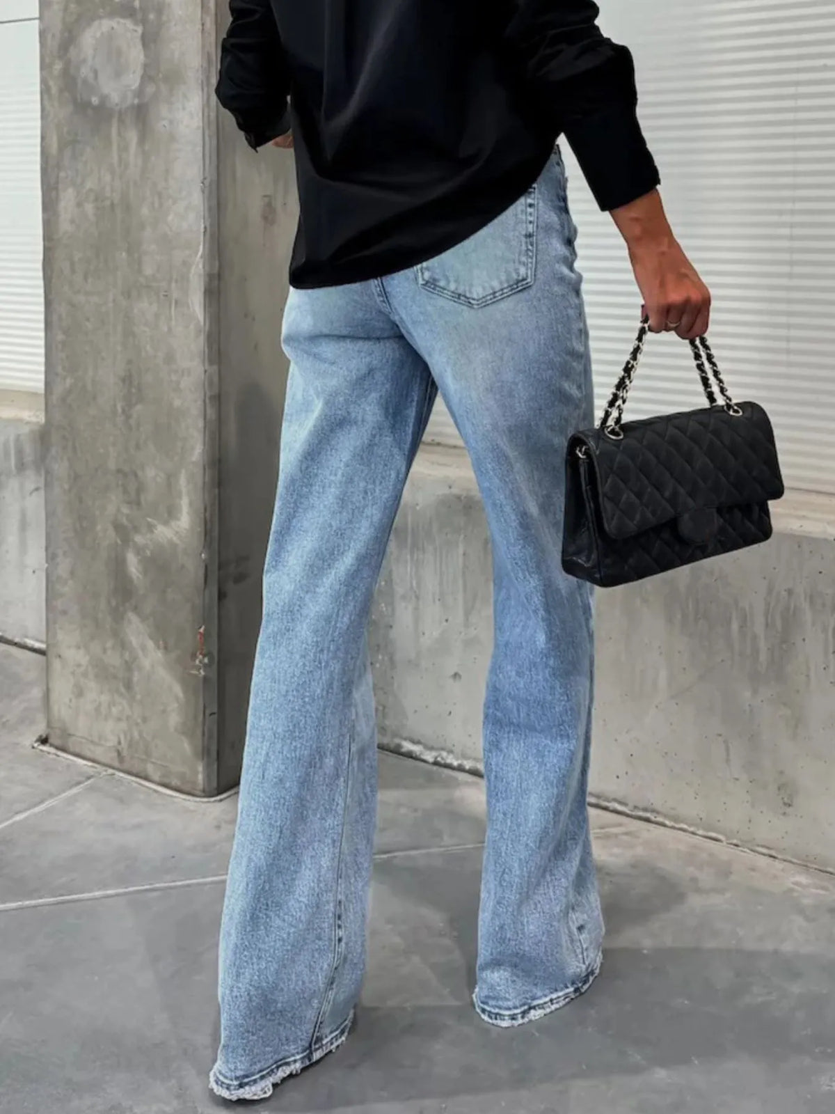 Person wearing light blue jeans and holding a black handbag against a concrete wall.