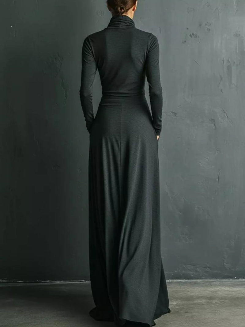 A woman viewed from behind wearing a long-sleeve, high-neck, floor-length black dress against a dark textured wall.