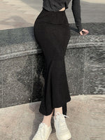Person wearing a black outfit with a slit skirt standing against a stone wall.