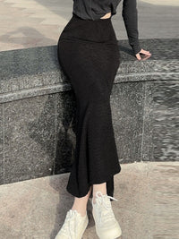 Person wearing a black outfit with a slit skirt standing against a stone wall.