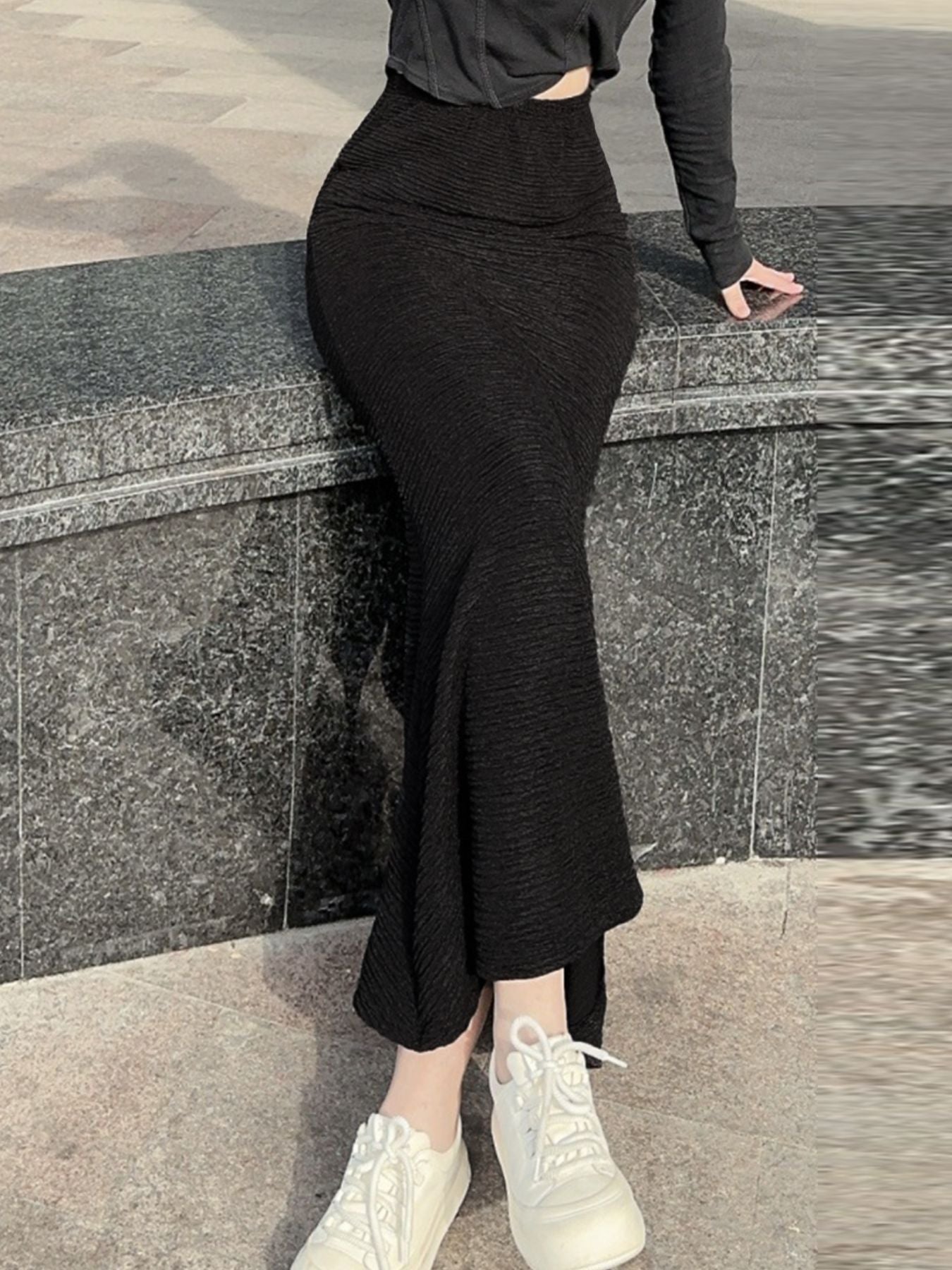 Person wearing a black outfit with a slit skirt standing against a stone wall.