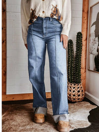 Person wearing blue jeans and a patterned shirt in a room with a cactus plant and framed picture.