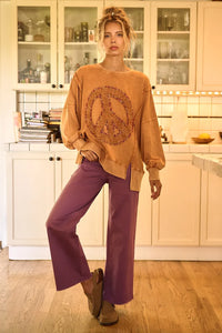 Woman wearing a beige sweater with a peace symbol and purple pants in a kitchen.