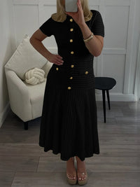 Person wearing a black dress with gold buttons in a room with a white chair and wooden floor.