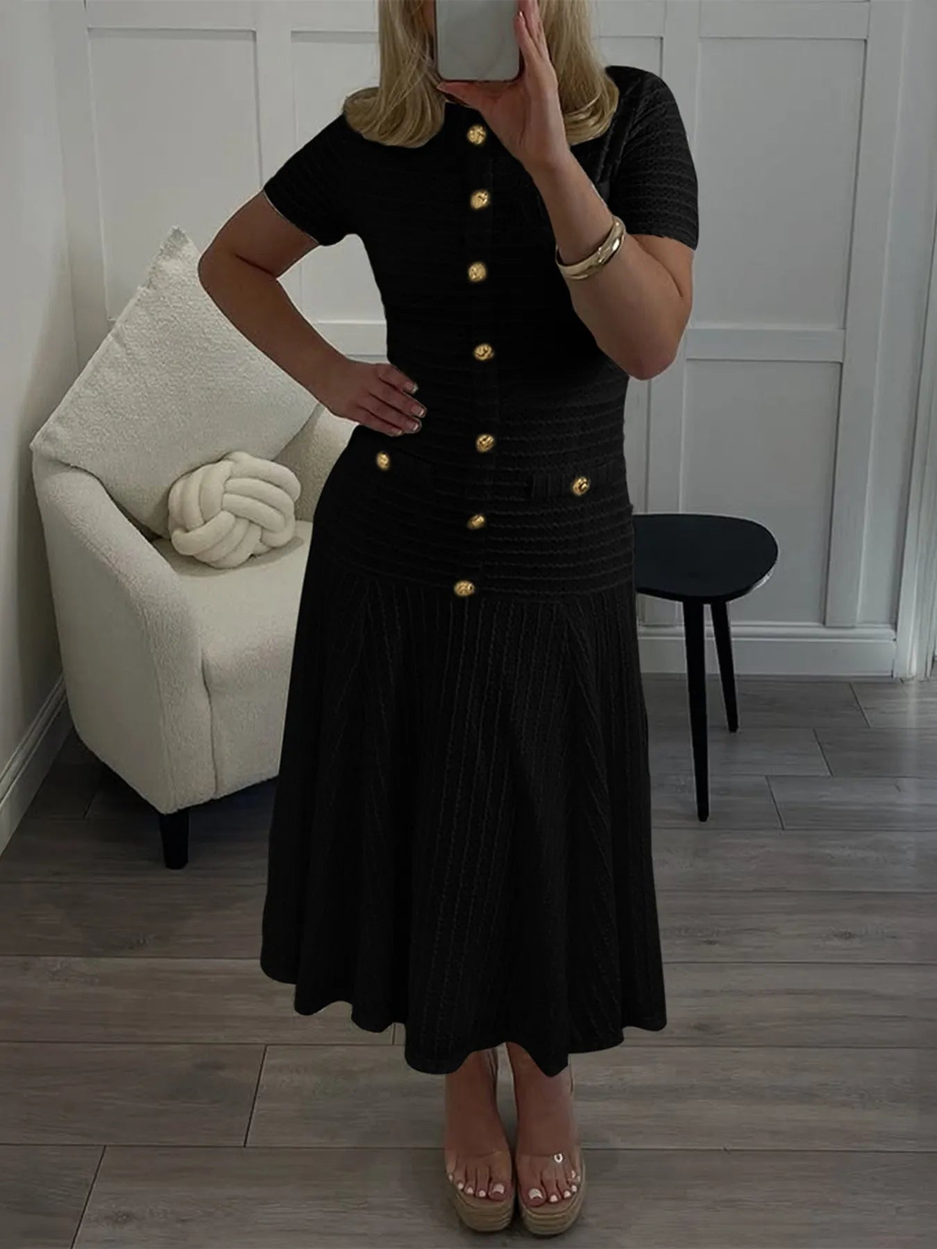 Person wearing a black dress with gold buttons in a room with a white chair and wooden floor.