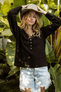 Woman wearing a black long-sleeve shirt and denim shorts standing among green plants.