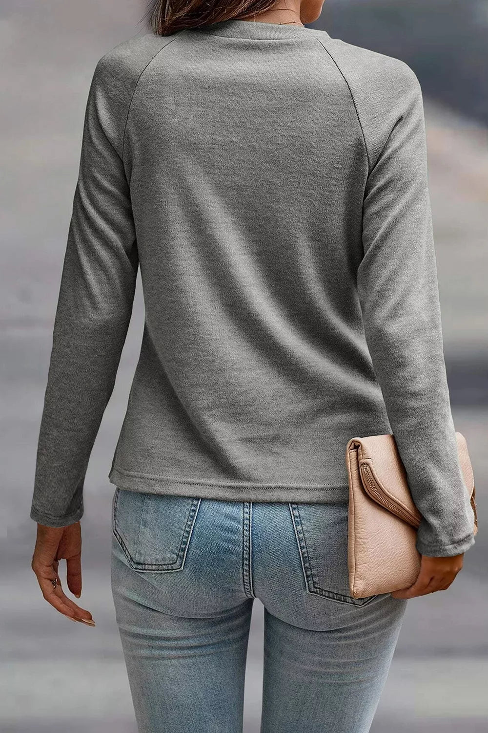 A woman shown from behind wearing a heather gray long-sleeve raglan top and light blue jeans, holding a beige clutch bag.