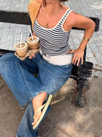 Woman sitting on a bench holding coffee cups, wearing a striped tank top and jeans.
