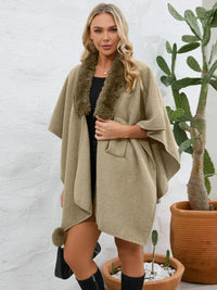 Woman wearing a beige poncho with fur trim standing in front of a white wall and potted plants.