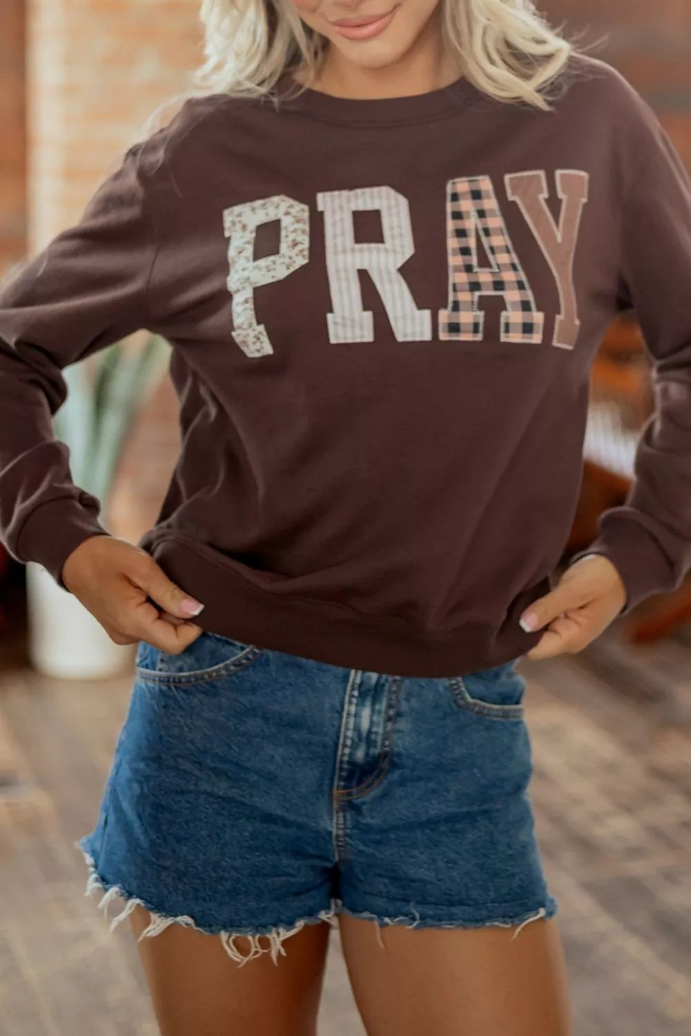 A woman wearing a brown long-sleeve crewneck sweatshirt with the word 'PRAY' printed across the chest in mixed patterned lettering, paired with denim shorts.