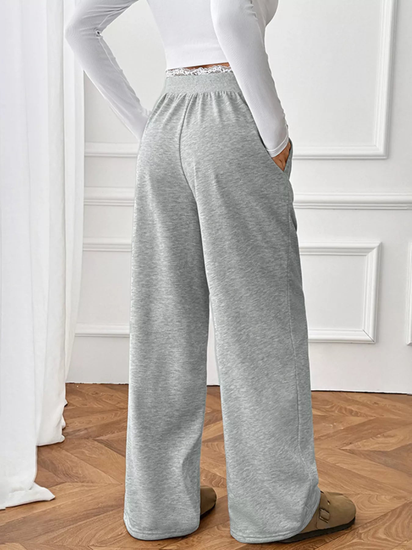 Person wearing gray sweatpants and a white long-sleeve top in a room with wooden flooring and white walls.