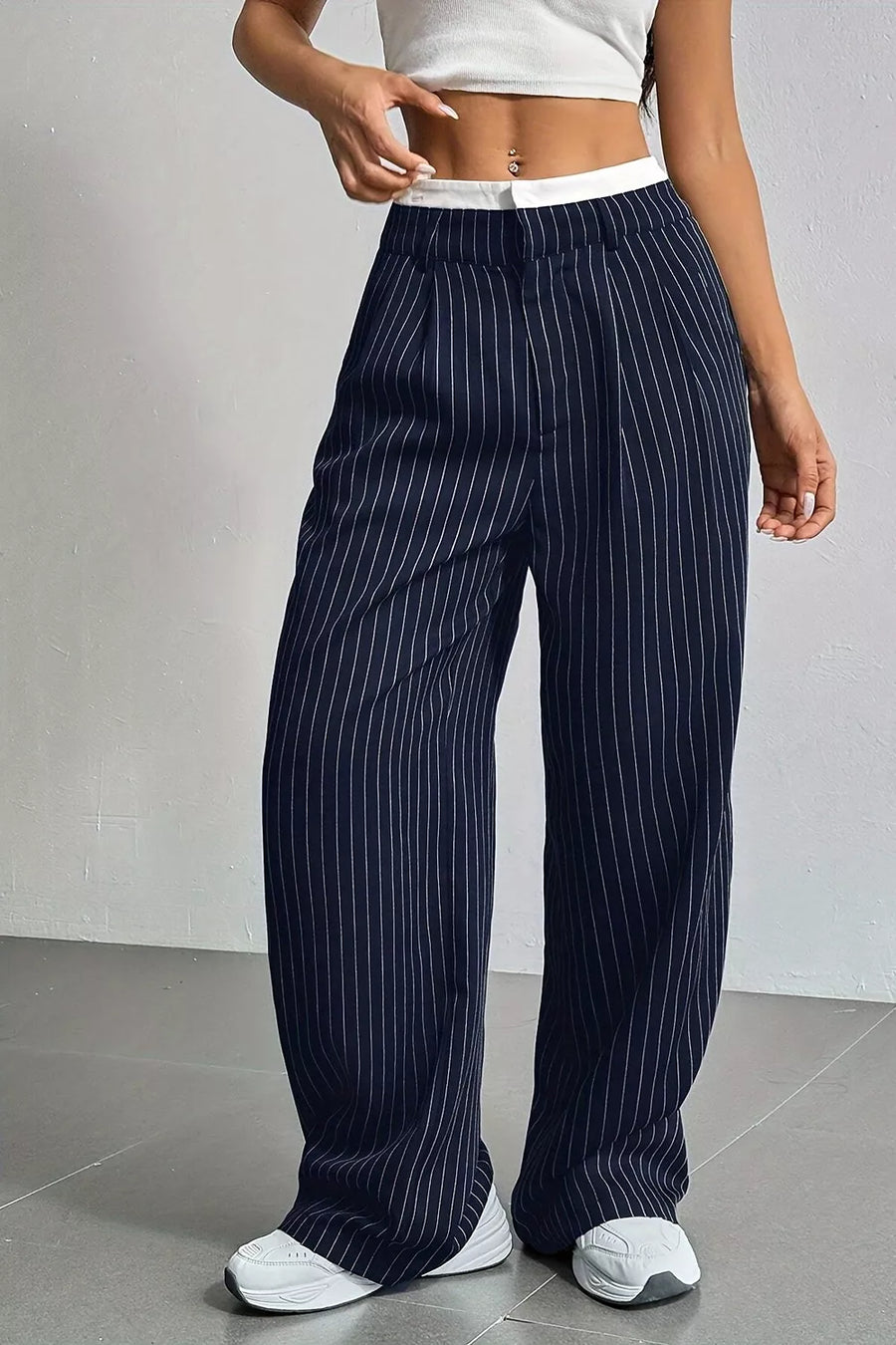 A person wearing navy blue wide-leg pinstripe pants with a white waistband, paired with a white crop top and white sneakers, standing against a light gray wall.