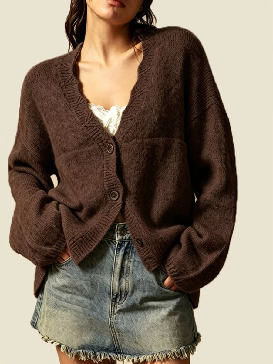 A woman wearing a dark brown chunky knit cardigan with a V-neck and button closure, paired with a white lace camisole and a light-wash denim mini skirt, set against a solid light beige background.