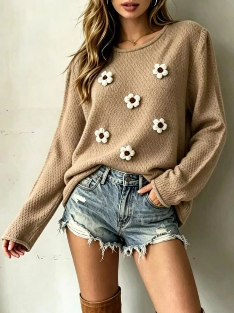 A woman wearing a beige textured knit sweater adorned with white daisy flower appliqués, paired with distressed denim shorts and brown boots, standing against a neutral wall.