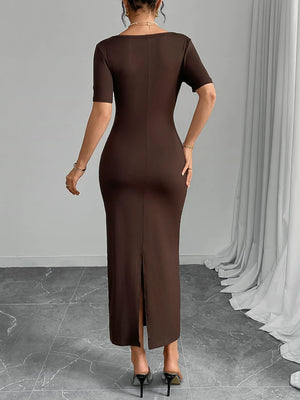 Woman wearing a brown dress with a slit, standing against a gray wall with white curtains.