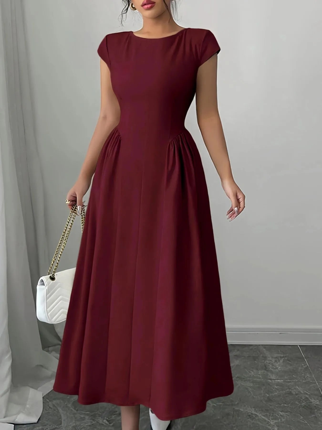 Woman wearing a burgundy dress holding a white handbag against a gray background