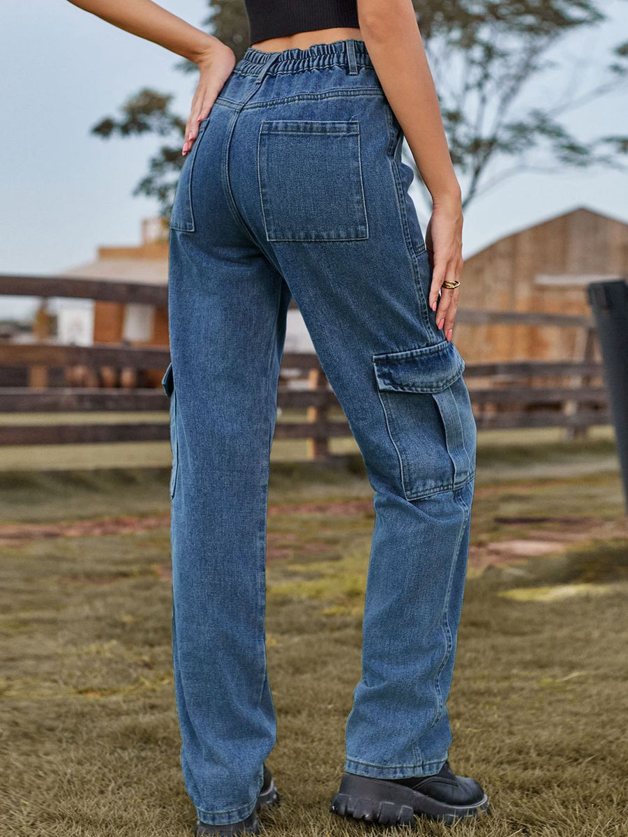 Person wearing blue cargo pants standing in a grassy field with a wooden fence and buildings in the background.
