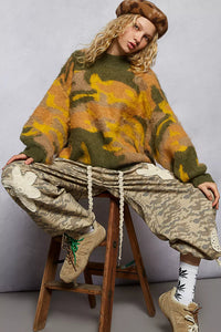 Person wearing a patterned sweater and baggy pants sitting on a stool against a plain background