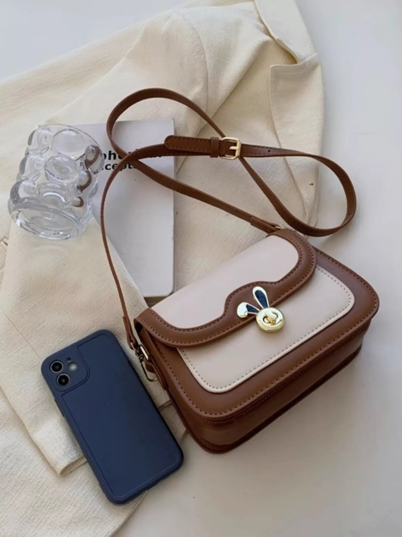 Small brown and beige handbag with a decorative clasp next to a blue phone on a light fabric background.