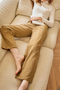 Person wearing beige pants and a white top, sitting on a beige couch holding a book.