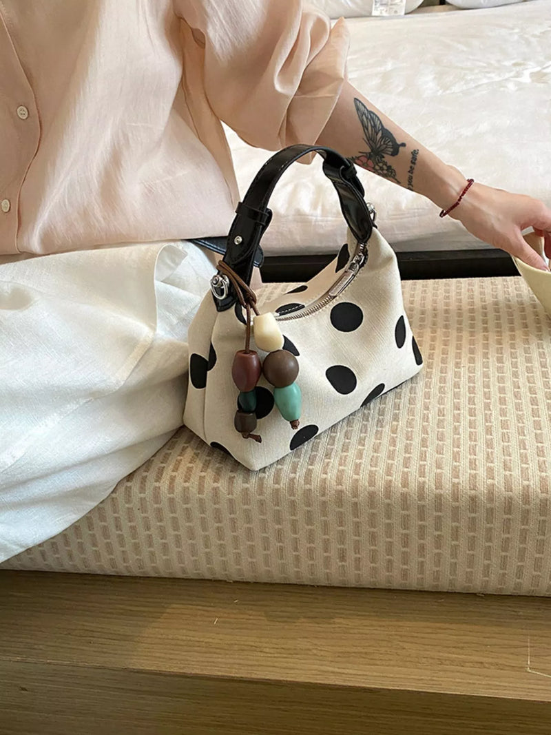 A cream-colored fabric handbag with black polka dots and a black leather handle, styled on a woven bench next to a person wearing a light pink blouse.