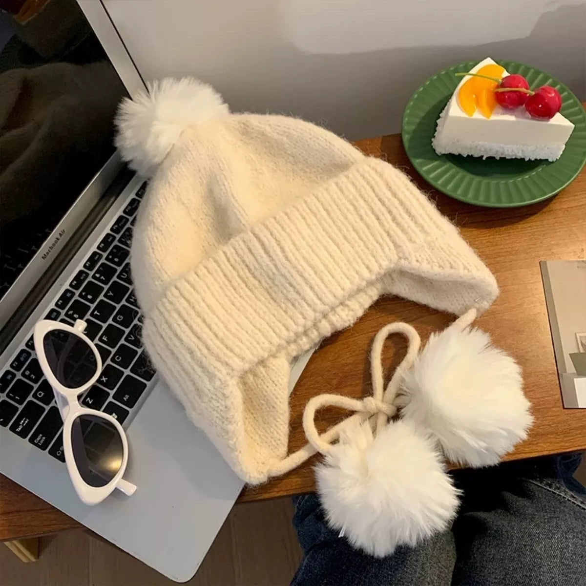 White knit beanie with pom-poms on a laptop next to a plate of cake.