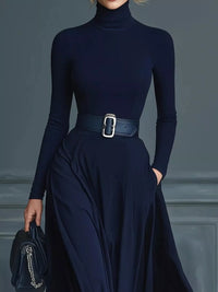 Person wearing a navy blue turtleneck and skirt with a belt, holding a matching handbag against a gray background.